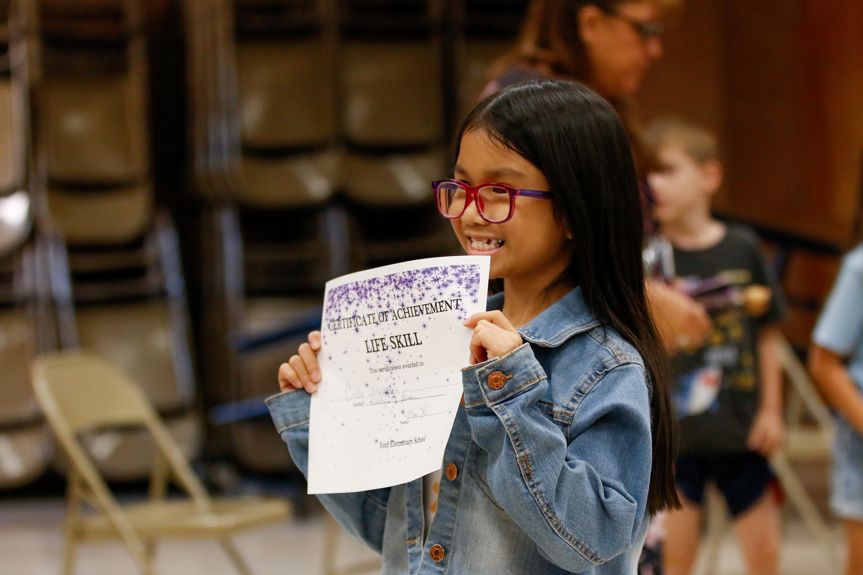 A little girl with dark hair and glasses smiles with her Life Skill certificate