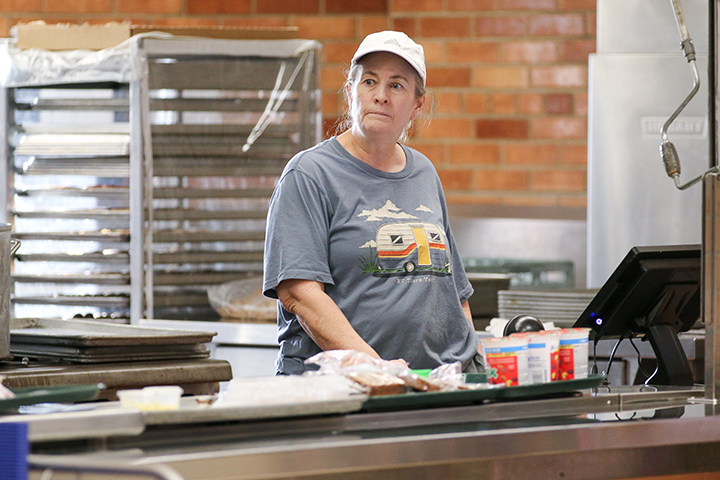 A woman stands in the cafeteria behind the counter, waiting to serve students