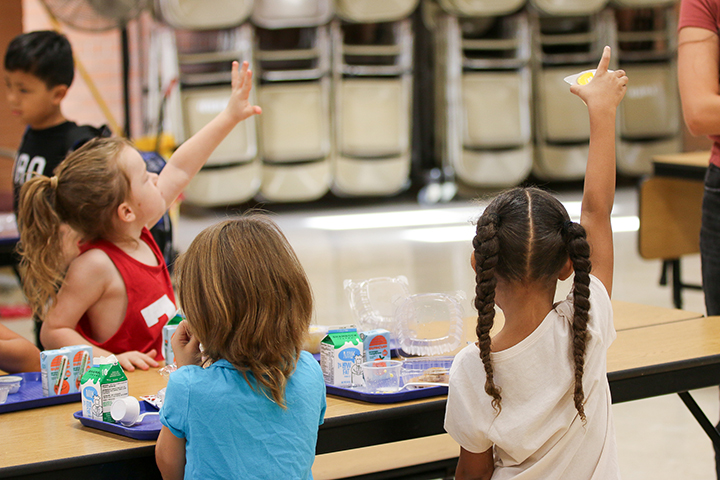 Two girls sitting at a table in the cafeteria raise their hands