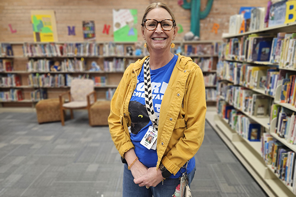 A woman in a yellow jacket, blue shirt and glasses smiles in the library