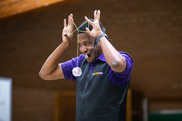 A man in a purple shirt and black vest does a yo-yo trick in front of his face