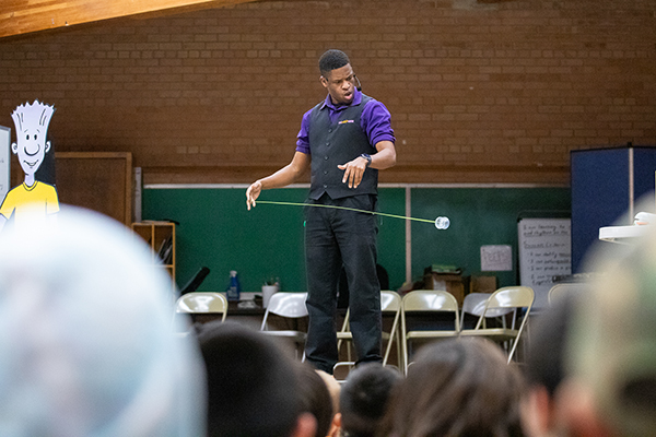 A man in a purple shirt and black vest does a yo-yo trick